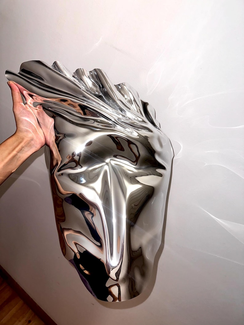 May include: A silver abstract sculpture with a smooth, reflective surface. The sculpture is shaped like a face with a long, pointed nose and a wide mouth.