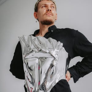 May include: A person wearing a black shirt with a silver, reflective, abstract, sculptural piece of body armor.