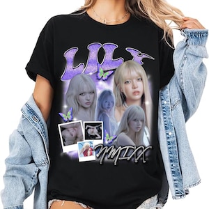 May include: Black t-shirt featuring a graphic design with the name "LILY" in purple glitter letters, images of a woman, butterflies, and the text "NMIXX". The shirt has short sleeves and a crew neck.
