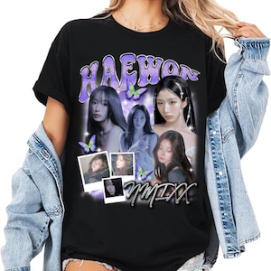 May include: Black t-shirt featuring a graphic design with the name "HAEWON" in purple, along with images of several women and butterflies. The word "NMIXX" is also visible in a stylized font. The shirt is worn with a denim jacket.