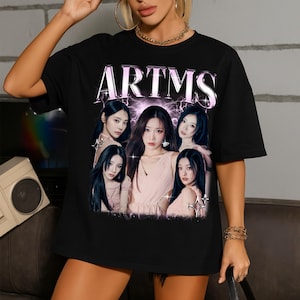 May include: Black t-shirt featuring the word "ARTMS" in a light purple, stylized font above a graphic of six women. The women are wearing pink dresses and have long, dark hair. The background has a lightning bolt design.