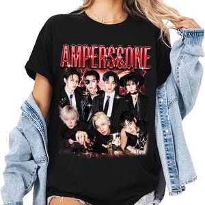 May include: Black t-shirt featuring a group of people with the text "AMPERS&ONE" in red. The graphic includes a group of people in black suits and formal wear. The shirt is worn by a person wearing a denim jacket.