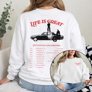May include: White sweatshirt with a graphic design on the back featuring a black car, figures, and the text "LIFE IS GREAT" and "JUST GOTTA BE A DICK SOMETIMES". Tour dates are listed below the graphic.