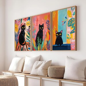 May include: Three framed art prints featuring black cats with colorful floral backgrounds. The cats are depicted in different poses, with one looking directly at the viewer. The prints are framed in natural wood.