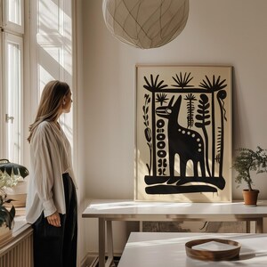 Op de afbeelding: Een zwart-witte illustratie van een gestileerd dier met een lange nek en een gepatroneerd lichaam, omringd door planten en bomen. De illustratie is ingelijst en hangt aan een muur in een kamer met een houten tafel en een persoon die bij een raam staat.