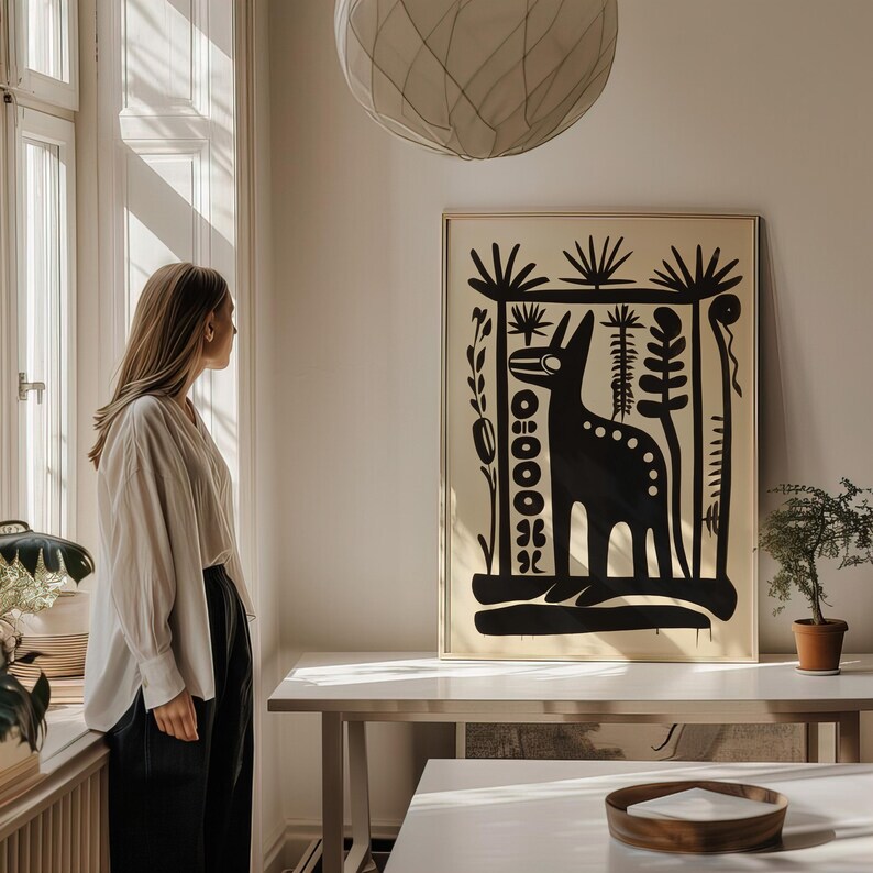 Op de afbeelding: Een zwart-witte illustratie van een gestileerd dier met een lange nek en een gepatroneerd lichaam, omringd door planten en bomen. De illustratie is ingelijst en hangt aan een muur in een kamer met een houten tafel en een persoon die bij een raam staat.