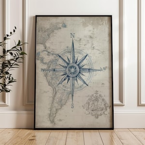 May include: Framed vintage map print with a navy blue compass rose. The map, in faded blue tones, depicts continents and oceans. A decorative ship illustration is in the lower right corner. The print is in a black frame, suitable for a home or office.