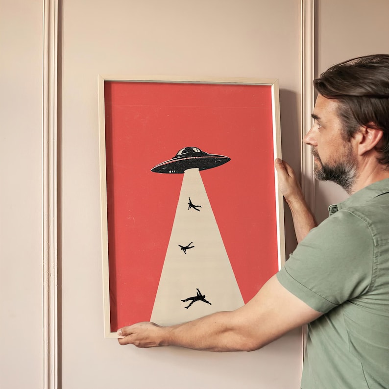 May include: Framed art print featuring a retro-style illustration of a UFO beaming up figures. The artwork has a red background with a black UFO and figures, and a white beam. The frame is a light wood color.
