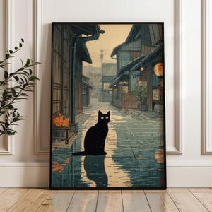 Puede incluir: Impresión artística enmarcada que representa un gato negro sentado en una calle adoquinada mojada en un pueblo japonés. La obra de arte presenta tonos apagados de azul, marrón y naranja, con arquitectura tradicional y linternas brillantes.