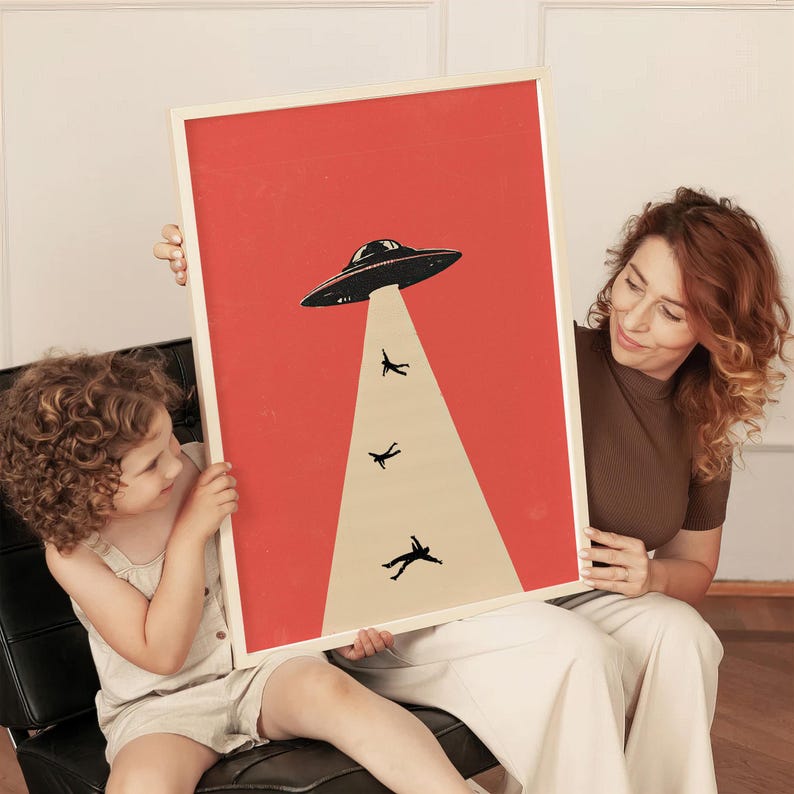 May include: A framed art print featuring a retro-style illustration of a black UFO beaming up silhouetted figures against a red background. The print has a cream-colored border and is held by two people.