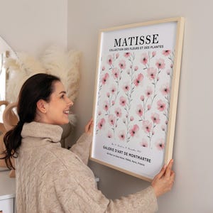 May include: A framed print of a watercolor painting of pink flowers with green stems on a white background. The print has the text "MATISSE COLLECTION DES FLEURS ET DES PLANTES GALERIE D'ART DE MONTMARTRE 83 Rue La Gare Nord, 75042, Paris, France" at the bottom.