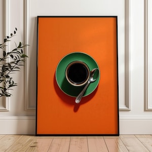 May include: A framed art print featuring a cup of black coffee on a green saucer with a spoon, set against an orange background. The artwork is displayed on a wooden floor, with a white wall and a plant in the background.