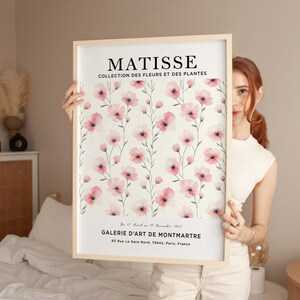May include: A framed art print featuring a watercolor painting of pink flowers with gray stems on a white background. The print has the text "MATISSE COLLECTION DES FLEURS ET DES PLANTES" at the top and "GALERIE D'ART DE MONTMARTRE 83 Rue Le Gare Nord, 75942, Paris, France" at the bottom.