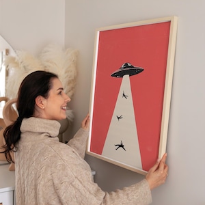 May include: Framed art print featuring a retro-style illustration of a black UFO beaming up three silhouetted figures against a red background. The print is in a light-colored wooden frame, perfect for adding a touch of whimsy to any space.
