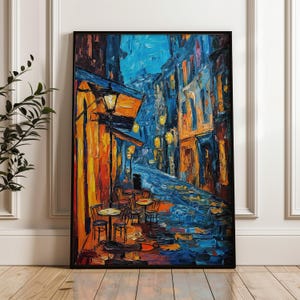 May include: A framed oil painting of a European street scene at night. The artwork uses a vibrant palette of blues, oranges, and yellows, with a focus on the reflections of streetlights on a wet cobblestone street. Tables and chairs are set up outside a cafe.