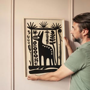 Op de afbeelding: Een ingelijste zwart-witprint van een gestileerd dier met een lange nek en vlekken, omringd door abstracte plantenvormen.