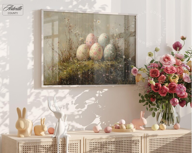 May include: A framed print featuring a still life of Easter eggs with floral patterns, set in a grassy area. The artwork is displayed above a wooden cabinet with decorative bunny figurines and a vase of pink roses. The image has the text "Arterville County".