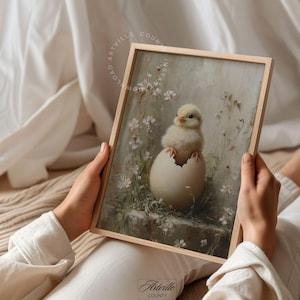 May include: A framed print featuring a yellow chick emerging from a cracked egg. The artwork has a soft, pastel color palette with floral accents and the text "LOVED ARTVILLE COUNTY INSTA". The frame is made of light-colored wood.