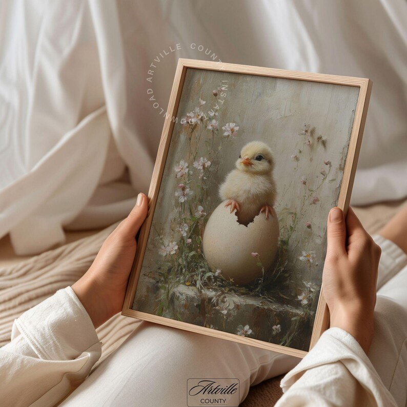 May include: A framed print featuring a yellow chick emerging from a cracked egg. The artwork has a soft, pastel color palette with floral accents and the text "LOVED ARTVILLE COUNTY INSTA". The frame is made of light-colored wood.