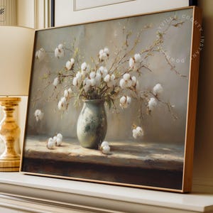 May include: A framed painting depicts a still life of cotton branches in a vase. The vase is a muted green, and the cotton bolls are white. The artwork has a soft, neutral color palette and is displayed on a white surface.