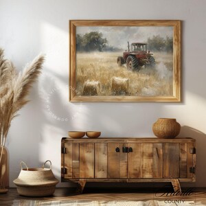 May include: A framed painting depicting a red tractor moving through a field of hay bales. The tractor is positioned in the centre of the field, with hay bales scattered around it. The painting is displayed against a white wall, with a wooden cabinet and a wicker basket in the foreground.