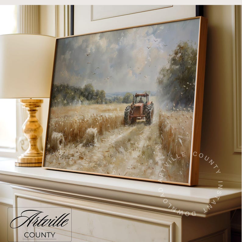 Puede incluir: Una pintura de paisaje enmarcada representa un tractor rojo en un campo de trigo bajo un cielo nublado. La obra de arte, con un marco de madera, presenta balas de heno y &aacute;rboles. La pintura est&aacute; apoyada en una superficie blanca, con el texto "Artville County" visible.
