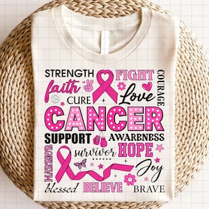 May include: Cream-colored t-shirt with a pink ribbon and the word "CANCER" in large, pink, polka-dotted letters. The shirt features words like "Strength," "Faith," "Cure," "Support," "Awareness," "Hope," and "Joy" in black and pink.