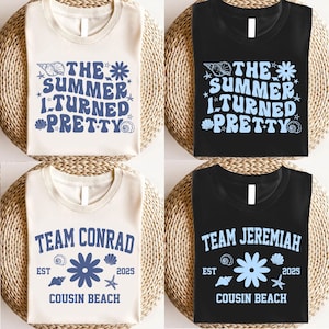 Op de afbeelding: Vier T-shirts met zomerse ontwerpen. Twee zijn crèmekleurig en twee zijn zwart. Elk shirt heeft blauwe tekst en afbeeldingen, waaronder schelpen, bloemen en de zinnen "The Summer Turned Pretty", "Team Conrad" en "Team Jeremiah".