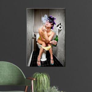 Op de afbeelding: Een ingelijste print toont een persoon in een gele badpak, zonnebril en krulspelden, die een sigaret rookt terwijl hij op een toilet zit. Een groene fles en een toiletpapierrol zijn ook zichtbaar. De badkamer heeft zwart-witte tegels.