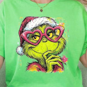 May include: A bright lime green t-shirt featuring a graphic illustration of a green creature wearing a red Santa hat and large pink heart-shaped glasses. The creature has a hand near its chin and is rendered in a colorful, artistic style.