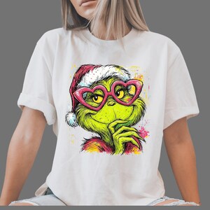 May include: A white short-sleeved t-shirt featuring a colorful graphic print of a green creature wearing a red and white Santa hat and large pink heart-shaped glasses. The illustration includes vibrant paint splatter accents in yellow, pink, and blue.