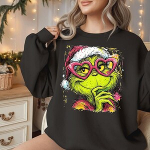 May include: A black long-sleeve sweatshirt featuring a graphic print of a green character wearing a red and white Santa hat and bright pink heart-shaped glasses. The illustration has a colorful, splattered paint effect around the edges.