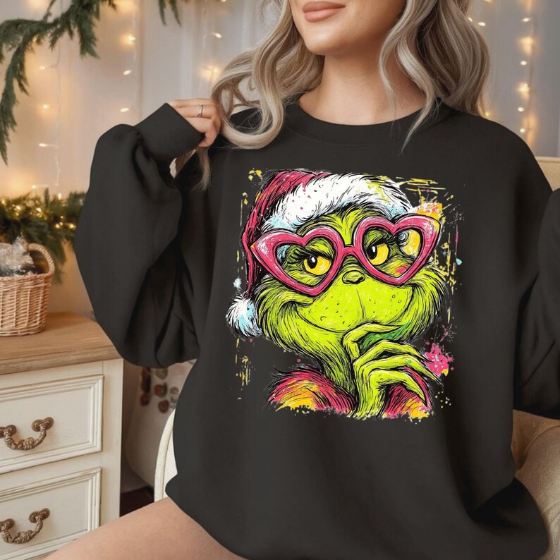 May include: A black long-sleeve sweatshirt featuring a graphic print of a green character wearing a red and white Santa hat and bright pink heart-shaped glasses. The illustration has a colorful, splattered paint effect around the edges.