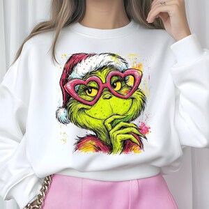 May include: A white long-sleeve sweatshirt featuring a graphic illustration of a green creature with yellow eyes, wearing a red and white Santa hat and pink heart-shaped glasses. The design includes colorful paint splatter accents.