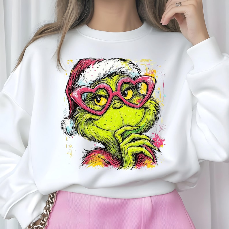May include: A white long-sleeve sweatshirt featuring a graphic illustration of a green creature with yellow eyes, wearing a red and white Santa hat and pink heart-shaped glasses. The design includes colorful paint splatter accents.
