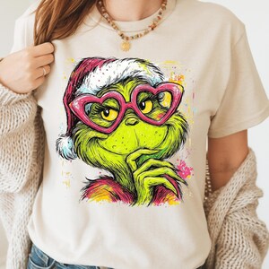 May include: A beige short-sleeve t-shirt featuring a colorful graphic print of a green creature wearing a red and white Santa hat and pink heart-shaped glasses. The illustration includes vibrant paint splatter accents in yellow, blue, and pink.