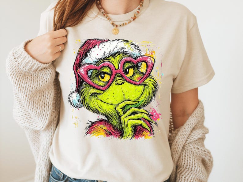 May include: A beige short-sleeve t-shirt featuring a colorful graphic print of a green creature wearing a red and white Santa hat and pink heart-shaped glasses. The illustration includes vibrant paint splatter accents in yellow, blue, and pink.