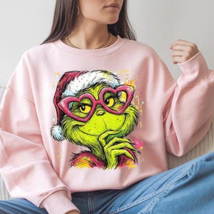 May include: A light pink crewneck sweatshirt featuring a graphic print of a green character wearing a red and white Santa hat and pink heart-shaped glasses. The character has a hand near its chin, with colorful paint splatter accents.