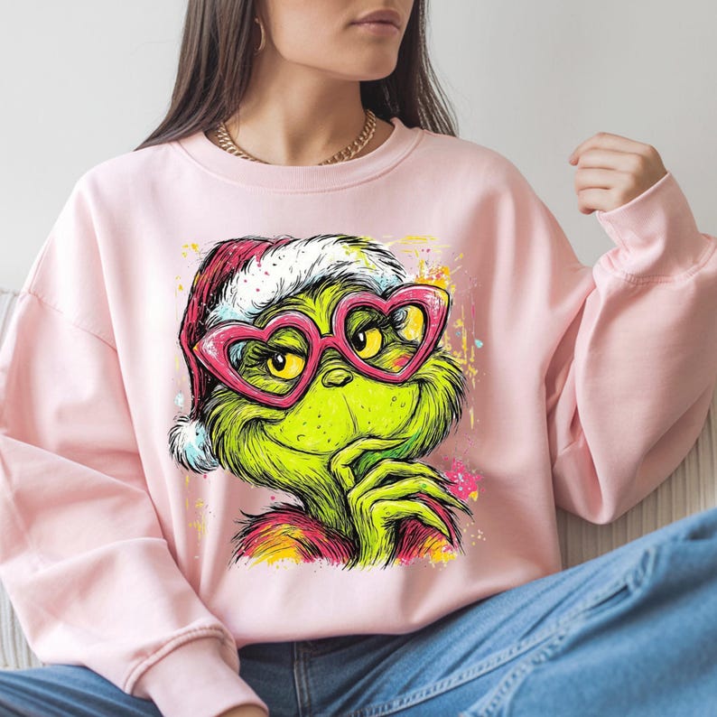 May include: A light pink crewneck sweatshirt featuring a graphic print of a green character wearing a red and white Santa hat and pink heart-shaped glasses. The character has a hand near its chin, with colorful paint splatter accents.