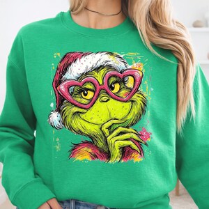 May include: A bright green crewneck sweatshirt featuring a graphic print of a green creature wearing a red and white Santa hat and large pink heart-shaped glasses. The illustration has a colorful paint splatter effect in yellow, pink, and orange.