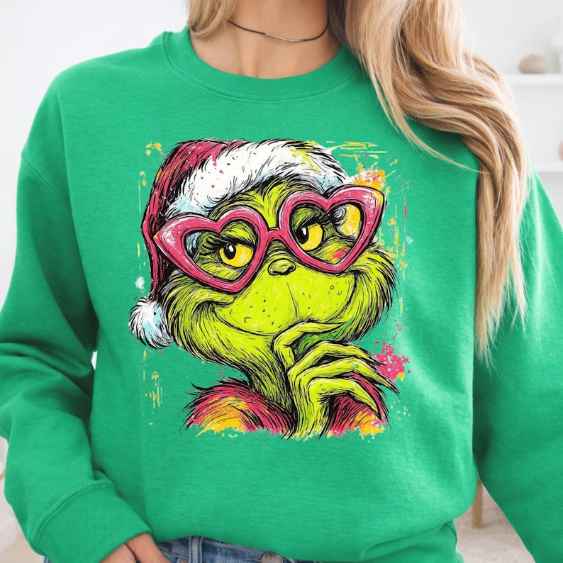 May include: A bright green crewneck sweatshirt featuring a graphic print of a green creature wearing a red and white Santa hat and large pink heart-shaped glasses. The illustration has a colorful paint splatter effect in yellow, pink, and orange.
