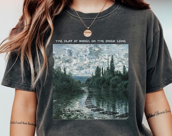 Può includere: T-shirt grigio scuro con una stampa del dipinto "The Play of Waves on the River Seine" di Claude Monet. L'opera raffigura una scena fluviale con una passerella in legno. Il testo "THE PLAY OF WAVES ON THE RIVER SEINE" è sopra.
