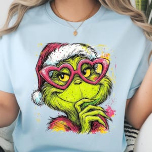 May include: A light blue t-shirt featuring a graphic illustration of a green, furry creature wearing a red and white Santa hat and large, pink, heart-shaped glasses. The design includes colorful paint splatters in yellow, pink, and red.