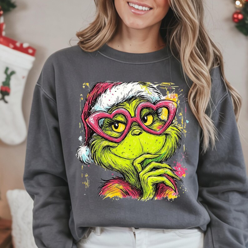 May include: A charcoal gray crewneck sweatshirt featuring a graphic print of a green creature wearing a red and white Santa hat and pink heart-shaped glasses. The illustration includes colorful paint splatter accents in yellow, pink, and blue.