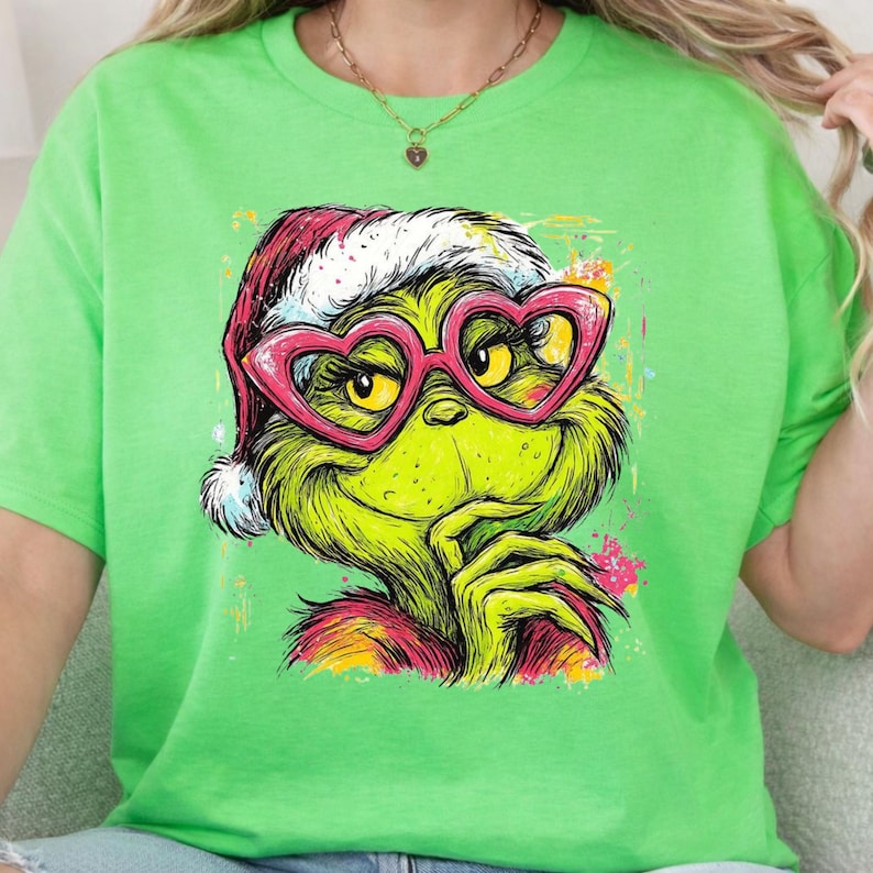 May include: A bright lime green t-shirt featuring a graphic illustration of a green creature wearing a red Santa hat and large pink heart-shaped glasses. The creature has a hand near its chin and is rendered in a colorful, artistic style.