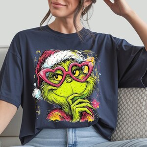 May include: A dark blue t-shirt featuring a graphic illustration of a green creature with yellow eyes wearing a red and white Santa hat and pink heart-shaped glasses. The design includes colorful paint splatter accents around the character.