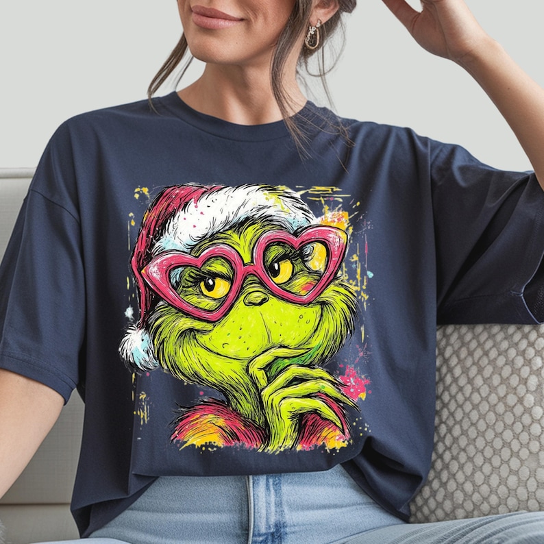 May include: A dark blue t-shirt featuring a graphic illustration of a green creature with yellow eyes wearing a red and white Santa hat and pink heart-shaped glasses. The design includes colorful paint splatter accents around the character.