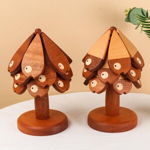 May include: Two wooden decorative objects shaped like stylized trees. Each tree has a round base, a stem, and a canopy made of layered, teardrop-shaped pieces. The pieces are a mix of light and dark brown wood, with white and black eye details.