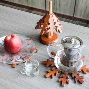May include: A wooden snowflake-shaped coaster with a glass teapot on top. A wooden Christmas tree-shaped coaster is in the background with a glass plate and a red apple on top.