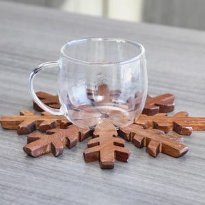 May include: A wooden snowflake coaster with a clear glass mug on top. The coaster is made of dark brown wood and has a detailed snowflake design.